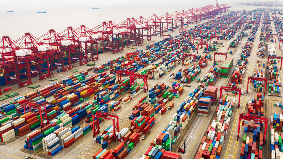 Aerial photo of a busy shipping port with rows of multicolored cargo containers, red cranes, and cargo ships docked in the harbor depicting global maritime logistics.